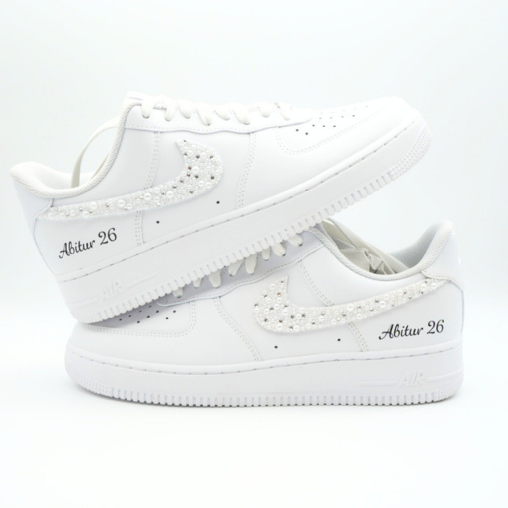 Weiße Nike Air Force 1 Sneaker mit perlenbesetztem Swoosh und seitlicher Aufschrift 'Abitur 26' in schwarzer Handschrift auf hellem Hintergrund.
