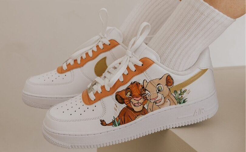 Weißer Nike Air Force 1 Custom Sneaker mit König-der-Löwen-Motiv, handgemalte Simba- und Nala-Figuren und oranger Swoosh, getragen mit weißen Sportsocken