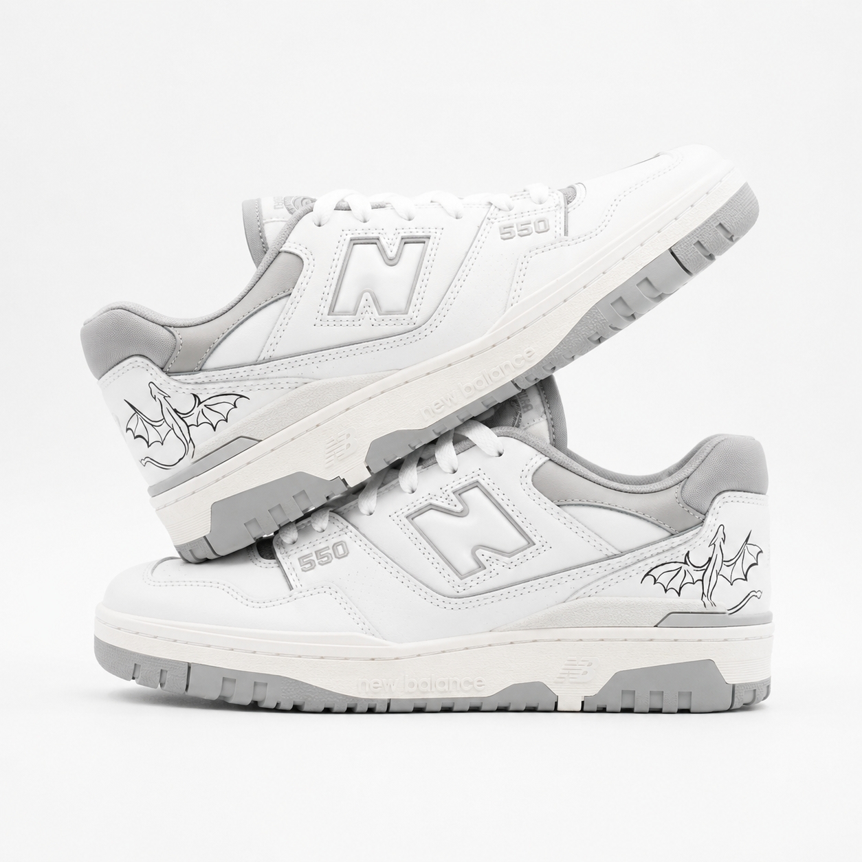 Paar weiße New Balance 550 Custom Sneaker mit grauen Akzenten und kleinen handgemalten Drachen-Linienzeichnungen auf den Seiten, gestapelte Präsentation