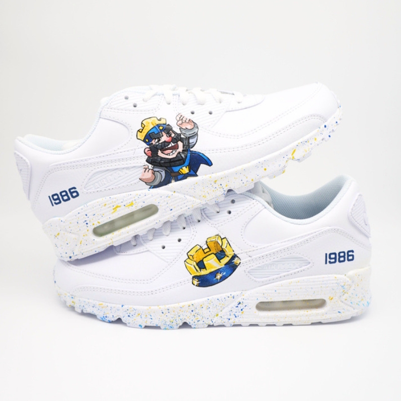 Paar weiße Nike Air Max Custom Sneaker mit Clash Royale Design, Königs-Figur Burg-Motiv Jahreszahl 1986 und blau-gelbe Farbspritzer auf der Sohle