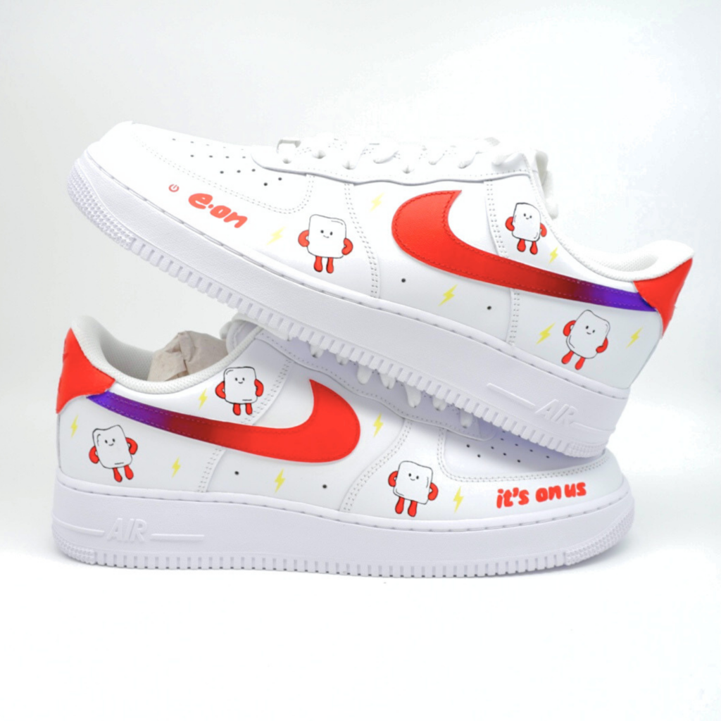 Weißer Sneaker im individuellen E.ON Branding mit lachenden Glühbirnen-Maskottchen, rotem Swoosh, lila Blitz-Streifen und Aufschrift 'it's on us' an der Ferse.