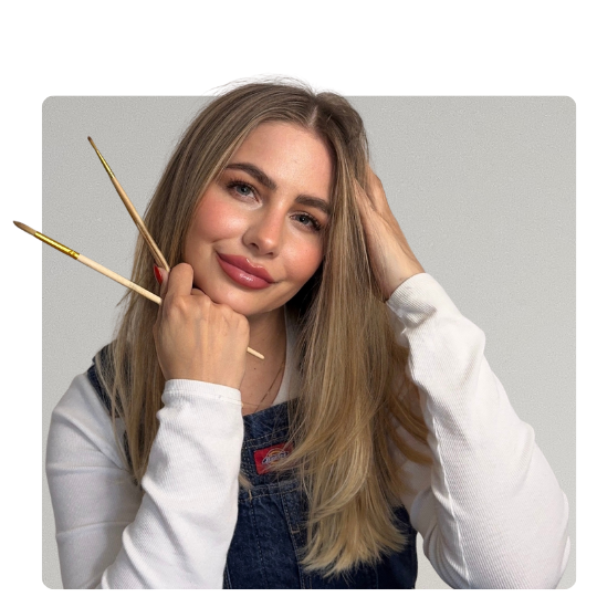 Porträtfoto von Kathi der Gründerin von unikartshoes, lächelnde junge Frau mit langen blonden Haaren hält zwei Pinsel in der Hand vor hellgrauem Hintergrund