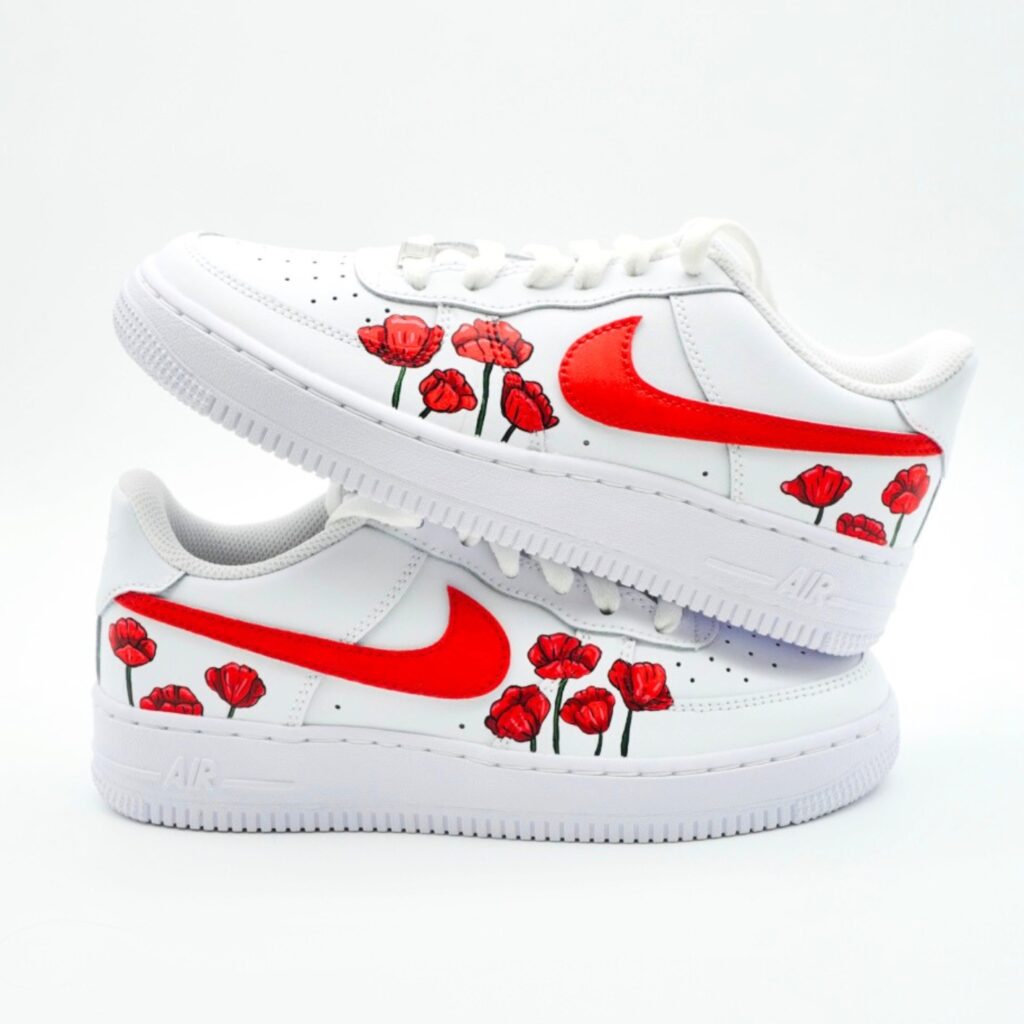 Weißer Nike Air Force 1 Sneaker mit handbemalten roten Mohnblumen entlang der Seiten und kräftig rot lackiertem Nike-Swoosh auf hellem Studiohintergrund.