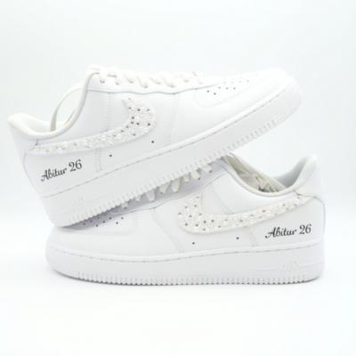 Weiße Nike Air Force 1 Sneaker mit perlenbesetztem Swoosh und seitlicher Aufschrift 'Abitur 26' in schwarzer Handschrift auf hellem Hintergrund.