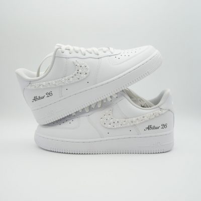 Zwei gestapelte weiße Nike Air Force 1 Sneaker mit silbrig glitzerndem Perlen-Swoosh und 'Abitur 26' Schriftzug auf hellem Hintergrund.