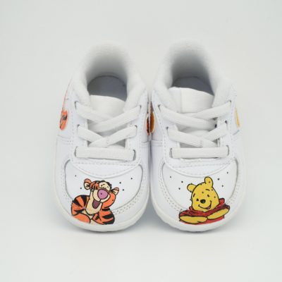 Weiße Babyschuhe mit handgemaltem Tigger- und Winnie Puuh Motiv an den Fersen, Schnürung und kleinem Honigtopf-Detail auf hellem Hintergrund.