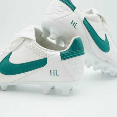 Paar weiße Fußballschuhe mit petrolfarbenem Swoosh und personalisierten Initialen HL, Stollensohle und gestapelte Präsentation vor hellgrauem Hintergrund