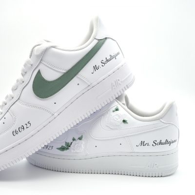 Zwei gestapelte weiße Nike Air Force 1 mit eukalyptus-grünem Swoosh, kleinen weißen Rosen, '06.09.25' und Schriftzügen 'Mr. Schultejans' und 'Mrs. Schultejans'.