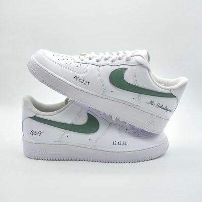 Zwei weiße Sneaker mit grünen Nike-Swooshs liegen gestapelt auf weißem Untergrund. Die Schuhe sind seitlich über der Zwischensohle mit individuellem Text, darunter Zahlen und Namen, versehen.