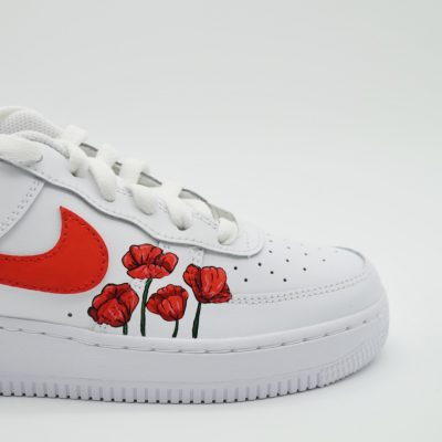 Ein weißer Sneaker mit einem leuchtend roten Nike-Swoosh und handgemalten roten Mohnblumen an der Seite, fotografiert vor einem schlichten hellen Hintergrund.