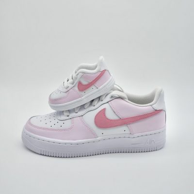 Vor einem schlichten Hintergrund sind zwei weiße Sneaker mit rosa Details zu sehen; ein kleiner Kinder-Sneaker liegt auf einem größeren Erwachsenen-Sneaker.
