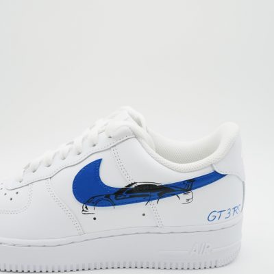 Ein weißer Nike Air Force 1-Sneaker verfügt über einen blauen Nike-Swoosh und eine detaillierte Illustration eines blau-schwarzen Sportwagens auf der Ferse mit der blauen Aufschrift „GT3 RS“ daneben.