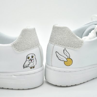 Ein Paar weiße Sneakers mit silbernem Glitzer an den Fersen. Ein Schuh hat ein Cartoon-Eulen-Design, der andere einen goldenen Ball mit Flügeln.