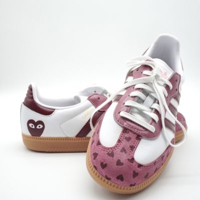 Ein Paar Turnschuhe mit weißen und kastanienbraunen Einsätzen, Gummisohlen, herzförmigen Mustern an der Spitze und einem Cartoon-Herz-Logo auf der Ferse, abgebildet auf weißem Hintergrund.