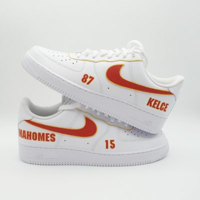 Weiße Sneaker mit Details der Kansas City Chiefs. Auf dem unteren Schuh ist hinten der Name des Spielers Mahomes und seine Trikotnummer 15 zu sehen, auf dem oberen Schuh ist hinten der Name des Spielers Kelce und seine Trikotnummer 87 zu sehen. Vor einem schlichten weißen Hintergrund liegt ein Schuh über dem anderen.