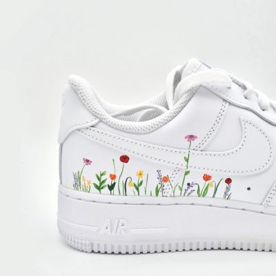 Weißer Nike Air Force 1 Sneaker mit einem handgemalten Design aus bunten Wildblumen und grünem Gras entlang der Seitensohle und der Ferse, vor einem schlichten weißen Hintergrund.