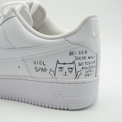 Nahaufnahme eines weißen Sneakers mit handgezeichnetem Katzen-Motiv, beidseitig erhobenen Pfoten und schwarzer Aufschrift 'Viel Spaß bei der Suche nach so tollen Kollegen wie uns'.