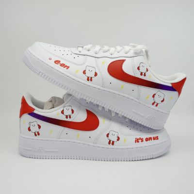Weißer Nike Air Force 1 Firmensneaker im E.ON Design mit rotem Swoosh, lila Blitz-Streifen, lachenden Glühbirnen-Maskottchen und Aufschrift 'it's on us' an der Ferse.