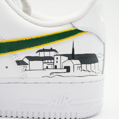 Nahaufnahme eines weißen Nike Air Force 1 mit handgemalter Stadtsilhouette in Schwarz-Weiß, Kirchturm und grün-gelben Banderolen entlang des Swoosh an der Ferse.