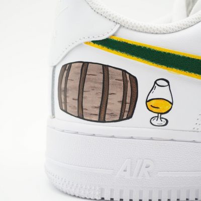 Nahaufnahme eines weißen Nike Air Force 1 mit handgemaltem Whisky-Fass, Glas mit goldener Spirituose und grün-gelber Banderole entlang des Swoosh an der Ferse.