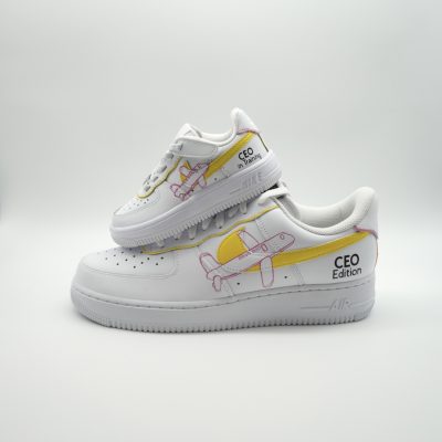Weißer Nike Air Force 1 Babysneaker mit gelbem Swoosh, hellrosa Mini-Flugzeug Illustrationen und 'CEO Edition' Schriftzug an der Ferse im hellen Studio-Setting.