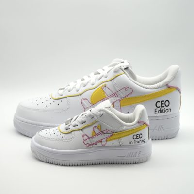 Zwei gestapelte weiße Nike Air Force 1 Babysneaker im 'CEO Edition' und 'CEO in Training' Design mit gelbem Swoosh und rosa Flugzeug-Motiven auf beiden Seiten.
