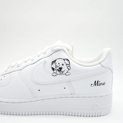 Nahaufnahme eines weißen Nike Air Force 1 mit fein gezeichnetem Dalmatiner-Welpen Porträt auf dem Swoosh und 'Mino' in schwarzer Schreibschrift an der Ferse.