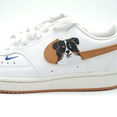 Nahaufnahme eines weißen Nike Sneakers mit gemaltem Border Collie Porträt und braunem Swoosh-Hintergrund auf hellem Mittelfeld mit blauem Mini-Swoosh.