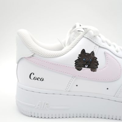 Nahaufnahme eines weißen Nike Air Force 1 mit rosa Swoosh, schwarzem Pomeranian-Hund Porträt und 'Coco' Schriftzug an der Ferse in dezenter Studioaufnahme.