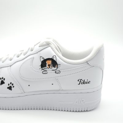 Nahaufnahme eines weißen Nike Air Force 1 mit handgemaltem dreifarbigen Katzen-Porträt, kleinen schwarzen Pfotenabdrücken und 'Pikie' Schriftzug an der Ferse.