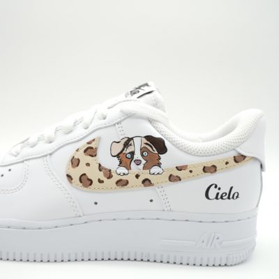 Nahaufnahme eines weißen Nike Air Force 1 mit Leopardenmuster Swoosh, kleinem Australian Shepherd Welpen Porträt und schwarzer 'Cielo' Schreibschrift an der Ferse.