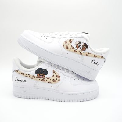 Zwei gestapelte weiße Nike Air Force 1 Sneaker mit Leopardenmuster Swoosh, Welpen-Porträts und personalisierten Namen 'Cielo' und 'Luana' an den Fersen.