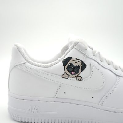 Nahaufnahme eines weißen Nike Air Force 1 mit handgemaltem Mops-Welpen Porträt auf dem Swoosh-Bereich vor neutralem Studio-Hintergrund.