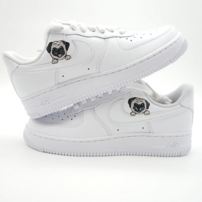 Zwei gestapelte weiße Nike Air Force 1 Sneaker mit kleinen handgemalten Mops-Porträts auf dem Swoosh-Bereich und schlichtem Look ohne weitere Akzente.
