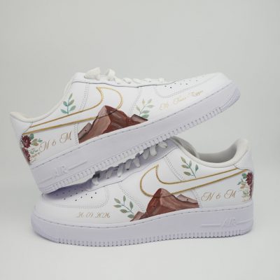 Zwei gestapelte weiße Nike Air Force 1 Hochzeitssneaker mit handgemaltem Berg-Motiv, goldenem Swoosh, Blumen-Ranken, Initialen 'M & M' und Datum '30.01.2026'.