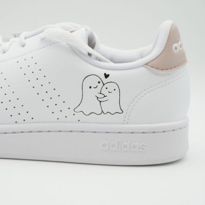 Nahaufnahme eines weißen Adidas Sneakers mit handgemaltem Geister-Pärchen, das sich liebevoll umarmt, kleinem schwarzen Herz und rosa Fersenkappe.