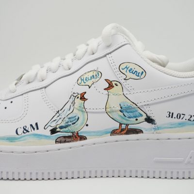 Nahaufnahme eines weißen Nike Air Force 1 mit handgemalten Möwen mit Sprechblasen 'Meins!', blau-gelben Wellen, Initialen 'C&M' und Datum '31.07.27' an der Ferse.