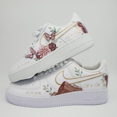 Zwei gestapelte weiße Nike Air Force 1 mit handgemaltem Berg-Motiv, Rosen, Blumen-Ranken, Schmetterlingen in Bordeaux-Rosa, Initialen 'M & M' und Datumsangaben.