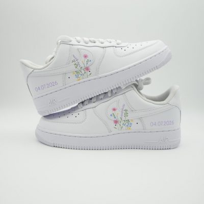 Zwei gestapelte weiße Nike Air Force 1 mit zarten pastellfarbenen Wildblumen-Sträußen, feinen Stielen und dezenter Datumsangabe '04.07.2026' an beiden Fersen.