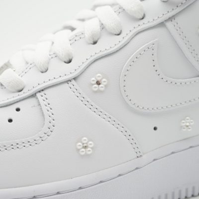 Nahaufnahme eines weißen Nike Air Force 1 mit aufgesetzten kleinen Perlenblumen mit goldener Mitte, dezent verteilt über die Seitenfläche im romantischen Hochzeitslook.
