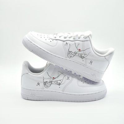 Zwei gestapelte weiße Nike Air Force 1 mit handgemaltem 'Pinky Promise' Finger-Motiv, kleinen roten Herzen und filigranen Initialen 'M' in feinem Strich.