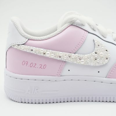 Nahaufnahme eines weißen Nike Air Force 1 Babysneakers mit hellrosa Akzenten, perlenbesetztem Swoosh und rosa Datumsangabe '07.02.20' an der Sohlenseite.