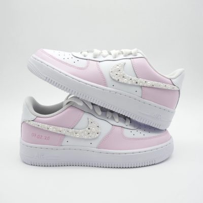 Zwei gestapelte weiße Nike Air Force 1 Babysneaker mit hellrosa Akzenten, perlenbesetztem Swoosh und feiner rosa Datumsangabe '07.02.20' an den Seiten.