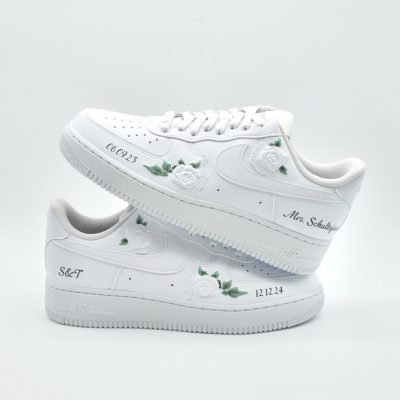 Zwei gestapelte weiße Nike Air Force 1 mit aufgesetzten weißen 3D-Rosen, grünen Blättern, Datum '06.09.25' und '12.12.24' sowie 'Mrs Schultej' und 'S&T' Schriftzügen.