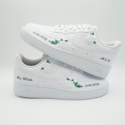 Zwei weiße Nike Air Force 1 Braut-Sneaker mit kleinen weißen Rosen, grünen Blättern, Schriftzug '13.06.2026', 'M & D' und 'Mrs Walde' in schwarzer Schreibschrift.