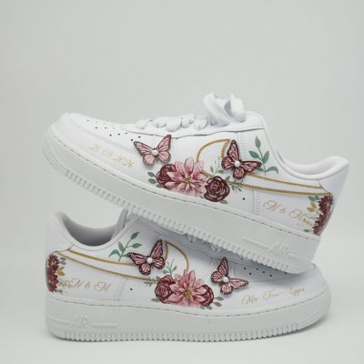 Zwei weiße Nike Air Force 1 mit Bordeaux-Rosa Blumen-Bouquet, aufgesetzten Schmetterlingen, goldenen Linien und Initialen 'M & M' im romantischen Hochzeitsdesign.