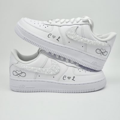 Zwei gestapelte weiße Nike Air Force 1 mit perlenbesetztem Swoosh, kleinen schwarzen Unendlichkeitszeichen, Herzen und schlichten Initialen 'C' und 'L' an den Fersen.