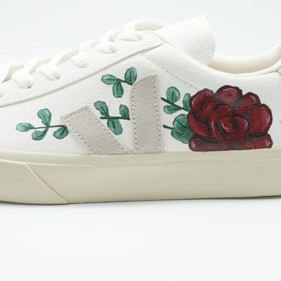 Nahaufnahme eines weißen Veja Sneakers mit großer handgemalter roter Rose, grünen Blättern und Eukalyptus-Ranken vor dem grauen Veja-Logo auf hellem Hintergrund.
