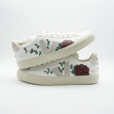 Zwei gestapelte weiße Veja Sneaker mit handgemalten roten Rosen, grünen Blättern und Eukalyptus-Ranken auf beiden Seitenflächen im romantischen Hochzeitslook.