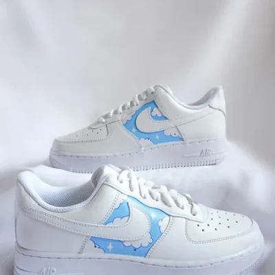 Zwei weiße Sneaker mit hellblauen Einsätzen, die Wolken und Sterne zeigen. Sie stehen auf weißem Stoffhintergrund und sind leicht seitlich ausgerichtet.
